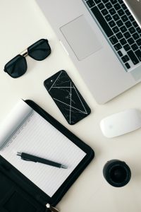 a desk with laptop, mouse, phone, sunglasses, camera lens, notepad and pen